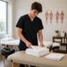 A male therapist in black medical scrubs folds a stack of white towels on a massage table in a bright, modern therapy room.