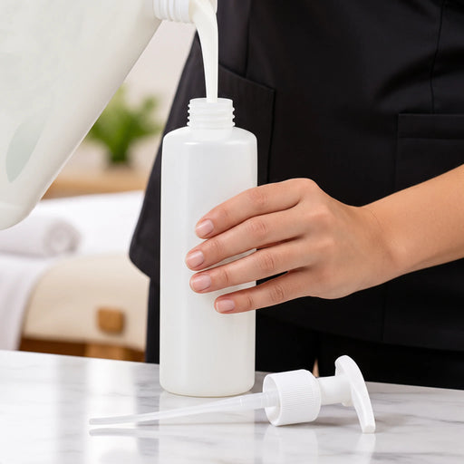 A person in a black uniform holds a BodyBest empty pump bottle while pouring a white liquid into it. The bottle has a matching white pump dispenser lying on a light marble countertop, with a softly blurred indoor background.