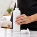 A person in a black uniform holds a BodyBest empty pump bottle while pouring a white liquid into it. The bottle has a matching white pump dispenser lying on a light marble countertop, with a softly blurred indoor background.