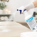 Gloved hand spraying Germiphene Genie Plus massage table cleaner onto a treatment table in a clean spa setting, with fine mist visible and towels and spa décor in the background.