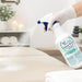Gloved hand spraying Myo-Ther Genie Plus massage table cleaner onto a treatment table in a clean spa setting, with fine mist visible and towels and spa décor in the background.