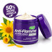 A purple jar of Nature's Kiss Anti-Flamme Extra herbal relief cream with a white cream swirl inside, shown with its lid leaning against it. Yellow flowers and green leaves decorate the base, and a circular badge in the top left reads '50% MORE ARNICA'.