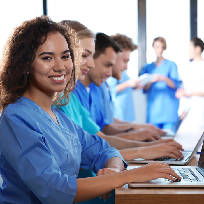 Massage therapy students in blue scrubs studying and working on laptops in a classroom.
