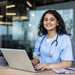 Massage therapy student in blue scrubs sitting at a desk and studying on a laptop in a classroom.
