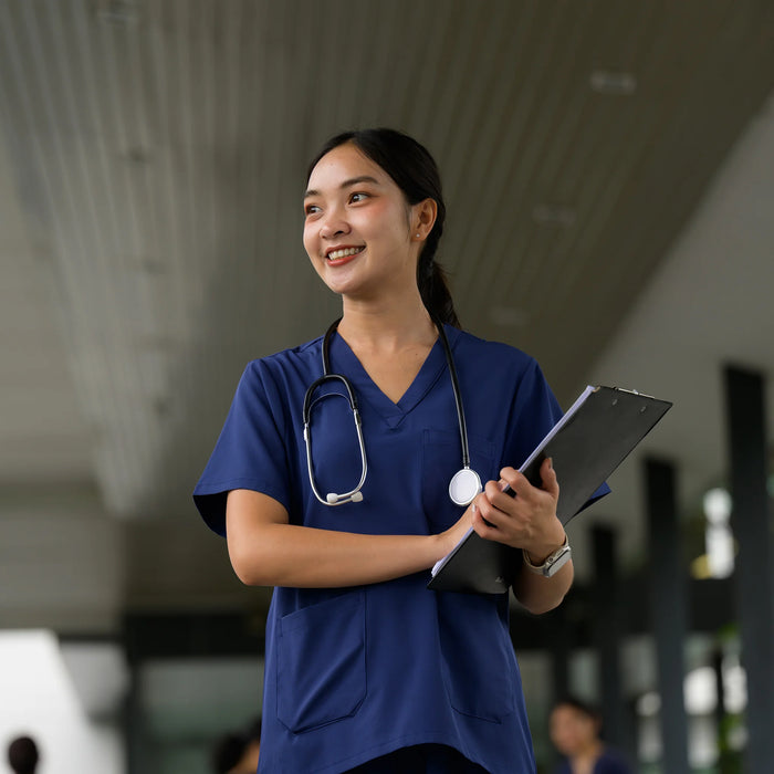 A smiling healthcare student in navy blue scrubs stands in a modern indoor corridor, holding a clipboard and wearing a stethoscope around her neck.
