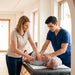 A lifestyle scene of a physical therapist and a student assisting an elderly man lying on a massage table in a bright, modern therapy room.