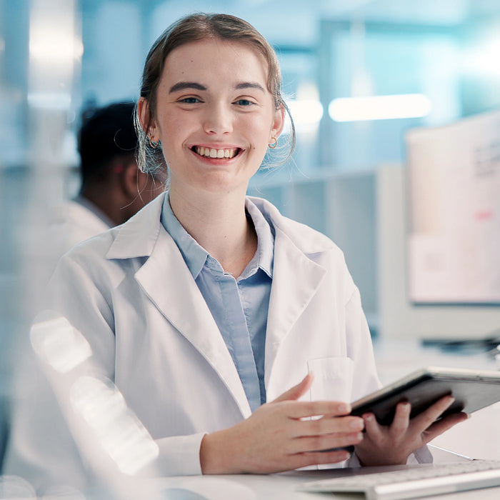 A smiling healthcare student in a white lab coat over a light blue shirt, holding a tablet in a bright, modern laboratory setting.