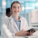 A smiling healthcare student in a white lab coat over a light blue shirt, holding a tablet in a bright, modern laboratory setting.