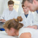 Massage students in white lab coats gathered around a patient lying on a table in a bright clinical setting.
