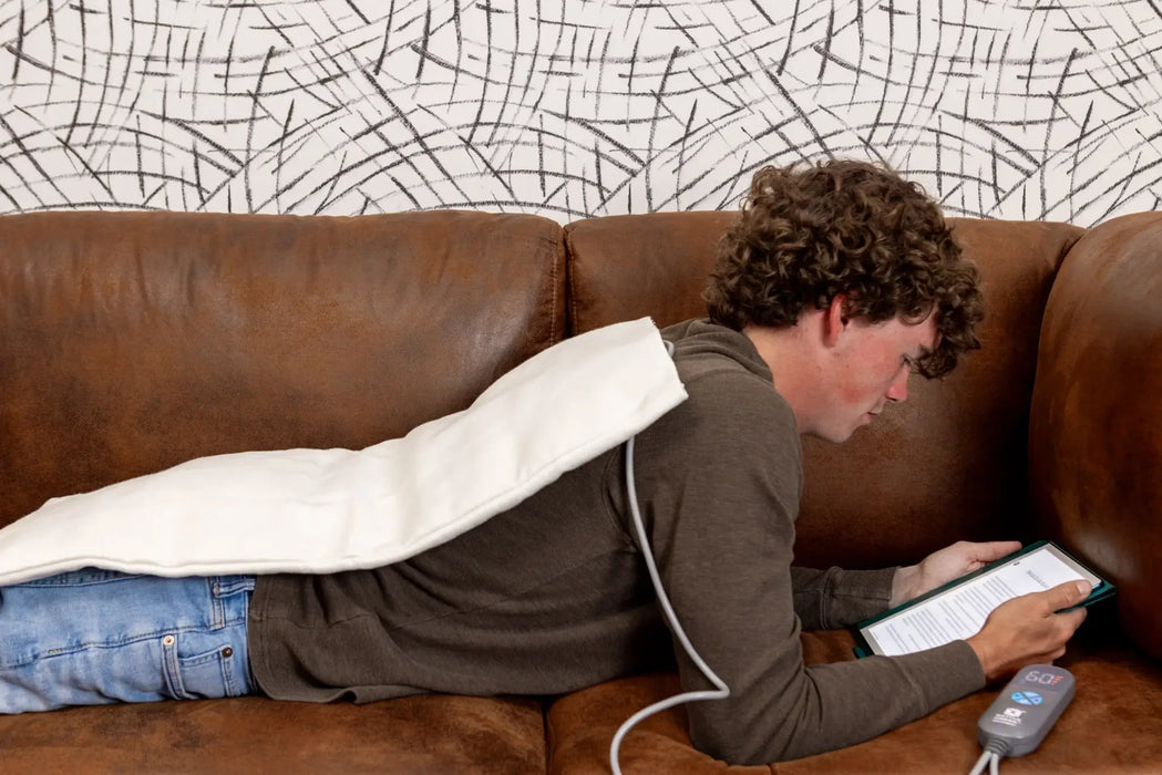 Person lying on a brown couch using a heating pad and tablet.